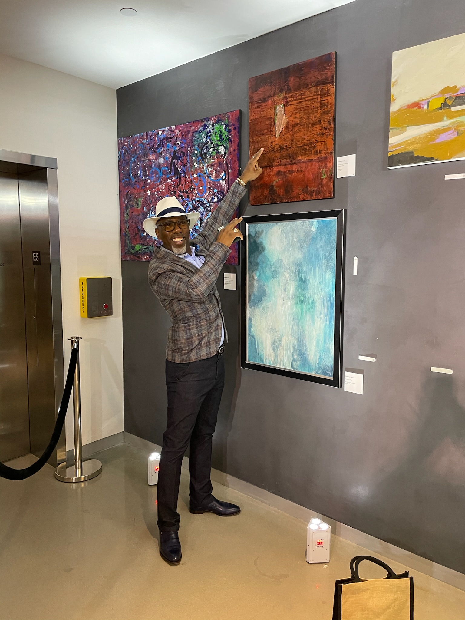 A collector in a panama hat points to a canvas on a dark gallery wall.